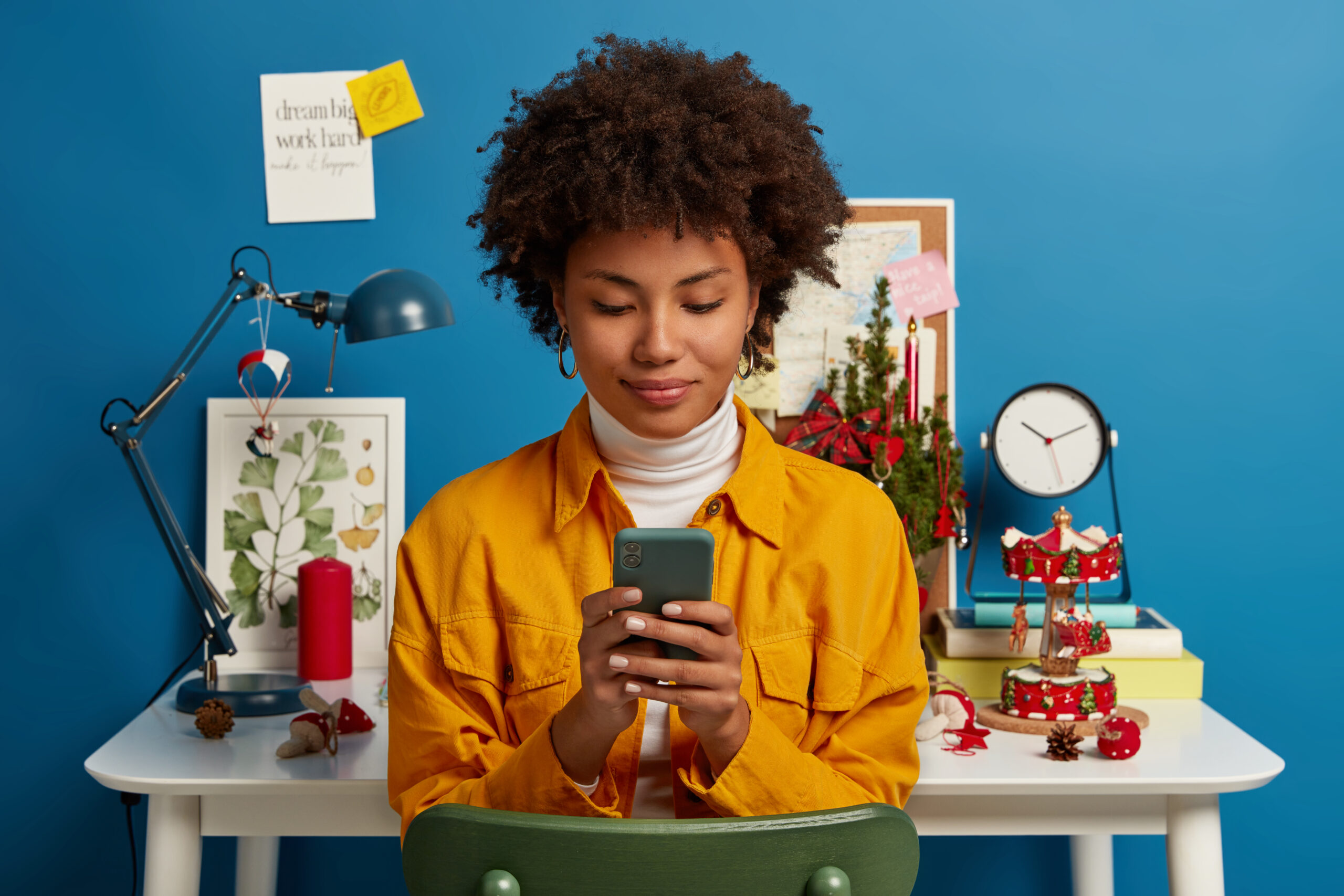 Pleased female student takes break from autodidact, uses cellular for chatting online, browses application, sends text message, checks mail via wifi, sits on chair near workplace, blue background Pleased female student takes break from autodidact, uses cellular for chatting online, browses application, sends text message, checks mail via wifi, sits on chair near workplace, blue background